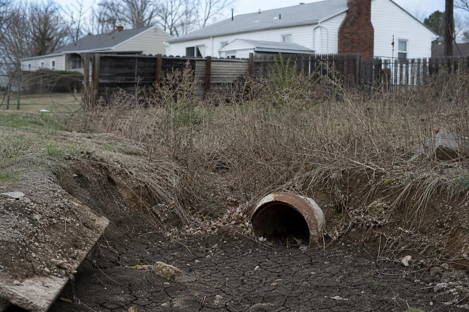 Eroded, overgrown drainage ditch with a rusted culvert pipe behind suburban homes